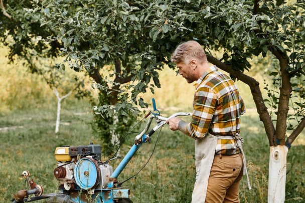 hombre barbudo guapo en traje casual con tatuajes usando cortacésped mientras está en el jardín en su granja - Foto, Imagen