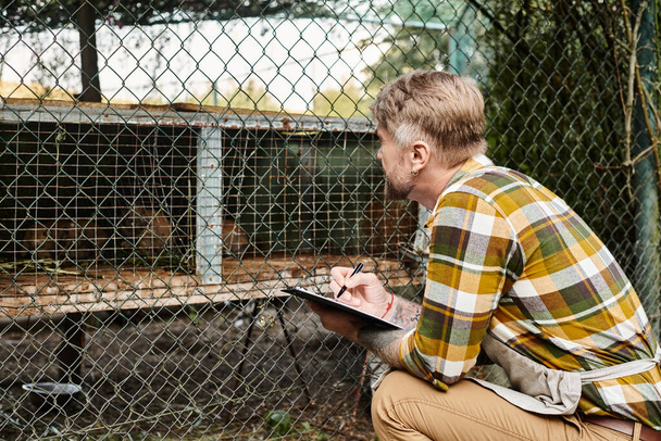 good looking bearded farmer in casual attire using clipboard to analyze aviary while in village - Photo, Image