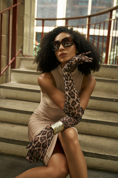 lockige schwarze Frau in Kleid, Handschuhen mit Animal-Print und Sonnenbrille, die auf Treppen in urbaner Umgebung sitzt - Foto, Bild