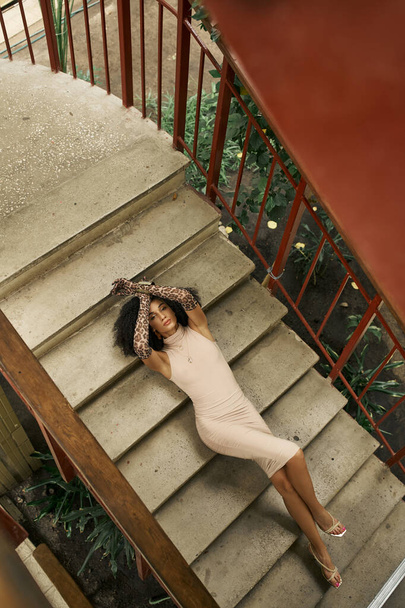 young black woman in dress and animal print gloves lying on stairs in urban garden, top view - Photo, Image
