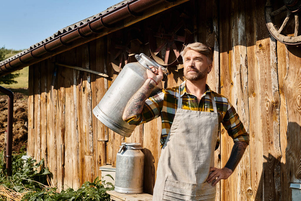 atraente barbudo homem em traje casual com tatuagens posando com churns de leite e olhando para longe, agricultor - Foto, Imagem