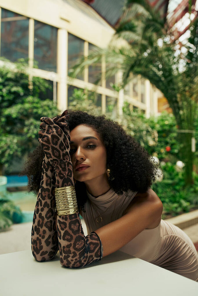 african american woman in animal print gloves and bracelets posing among plants in urban garden - Photo, Image