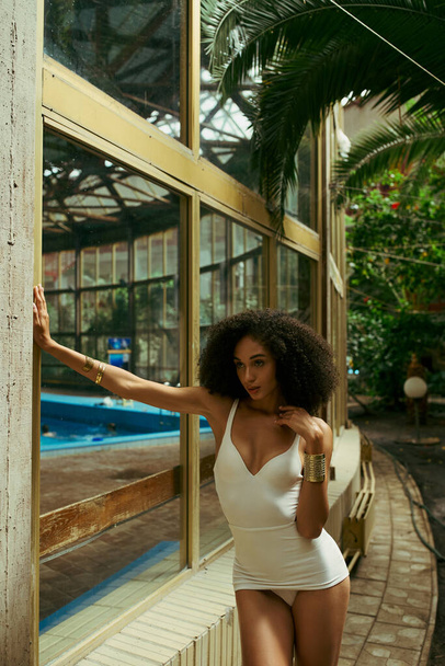 кучерява афроамериканка в купальнику позує в зеленому і екзотичному міському саду, різноманітність - Фото, зображення