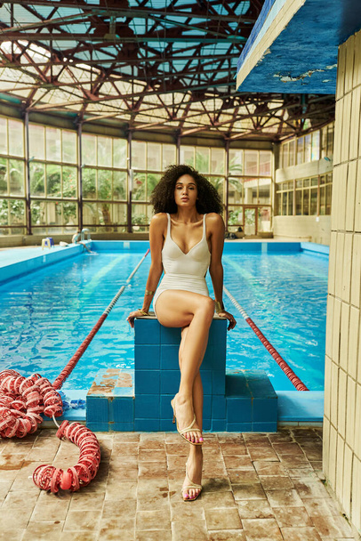 krullend en mooi Afrikaans amerikaanse vrouw in elegante badmode en hoge hakken zitten bij het zwembad - Foto, afbeelding