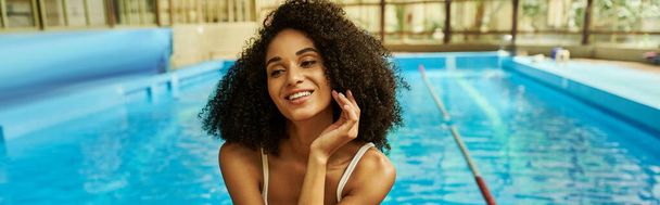 šťastný africký Američan žena v plavkách sedí u bazénu s modrou vodou, prapor - Fotografie, Obrázek