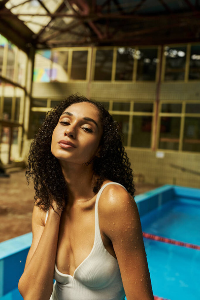 entspannte junge afrikanisch-amerikanische Frau mit nassen lockigen Haaren und Haut am Pool stehend, gelassen - Foto, Bild