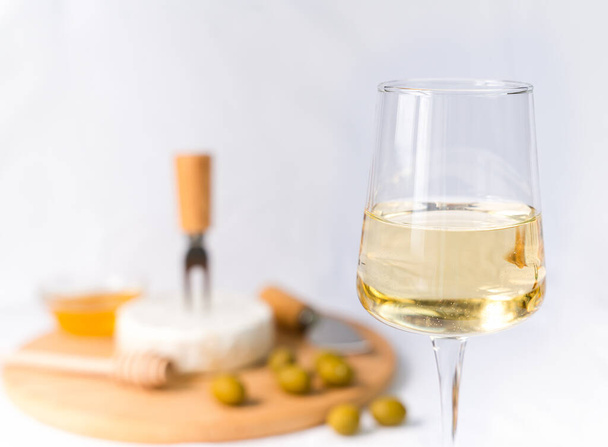 Ein Glas Weißwein und im Hintergrund eine Vorspeise aus Camembert und Oliven. Nahaufnahme. Selektiver Fokus. - Foto, Bild