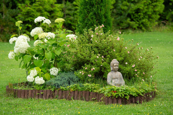 Meditierende Buddha-Figur sitzt in einer kleinen Insel aus Blumen, Potentilla-Büschen und Hortensien im Blumenbeet, umgeben von natürlicher Holzpalisade. - Foto, Bild