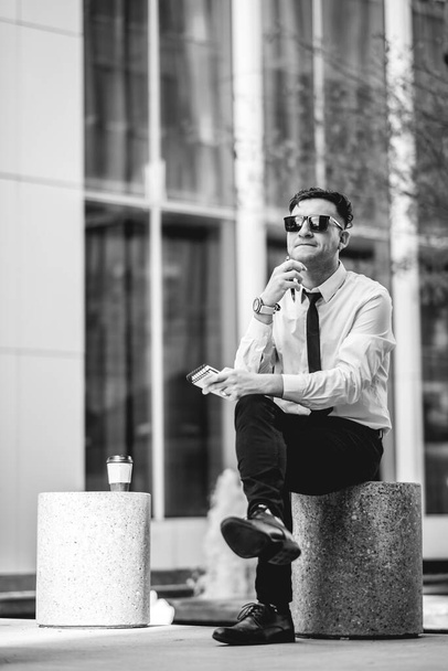 Professioneller, moderner und selbstbewusster junger Geschäftsmann mit weißem Hemd und schwarzer Krawatte, der ein Notizbuch und eine Kaffeetasse aus Papier im Stadtpark des Geschäftsviertels (in schwarz-weiß) in der Hand hält) - Foto, Bild