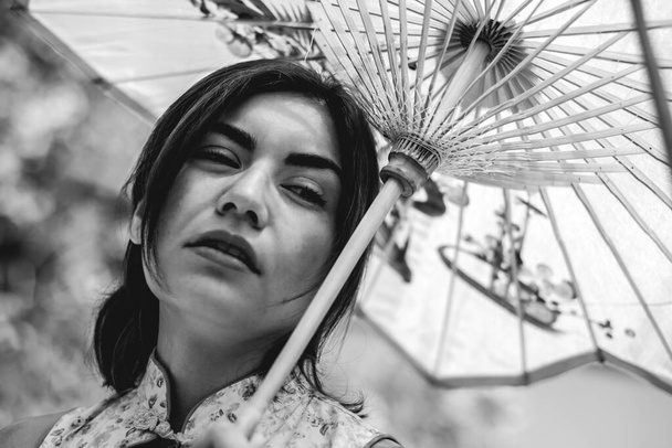 Impresionante belleza y estilo japonés: chica morena con tatuajes, vestido asiático y paraguas de papel, deleitándose en un día soleado con cielo despejado en un jardín japonés (en blanco y negro) - Foto, imagen
