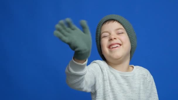 Szczęśliwe dziecko w dzianinie kapelusz i rękawiczki falujące hello na niebieskim tle - Materiał filmowy, wideo