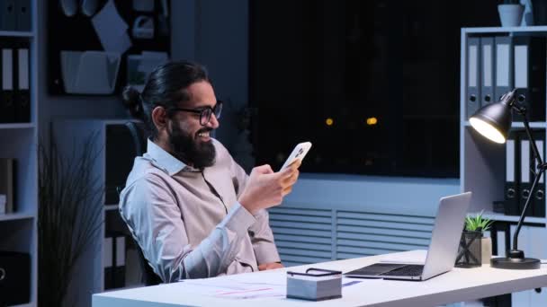 Ejecutivo indio hombre positivo utilizando el teléfono durante el descanso del trabajo y riendo en la oficina de la noche. Navegar por Internet, mensajería en las redes sociales, leer noticias. - Imágenes, Vídeo