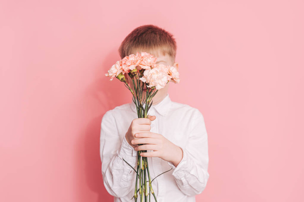 Garçon dans une chemise blanche couvre son visage avec un bouquet d'œillets, son visage devant un fond rose. - Photo, image