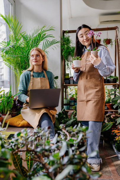 Függőleges lövés a bolt tulajdonosa középkorú szőke nő üzletasszony segítségével laptop dolgozik, ügyelve a növények pot neki koreai asszisztens segít csinál leltár hangulatos növény boltban.. - Fotó, kép