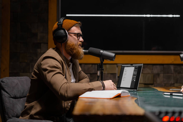 Un podcaster masculino entrevistando a un invitado en un estudio de grabación. Tipo barbudo sentado en la mesa con portátil y micrófono - Foto, imagen