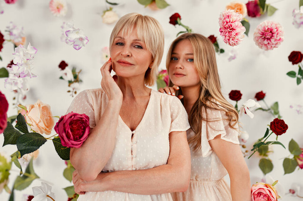 Feliz Día de la Madre y Día de la Mujer. Mamá y su hija en vestidos de pie aislados sobre un fondo blanco, hay muchas flores en el aire alrededor. - Foto, imagen