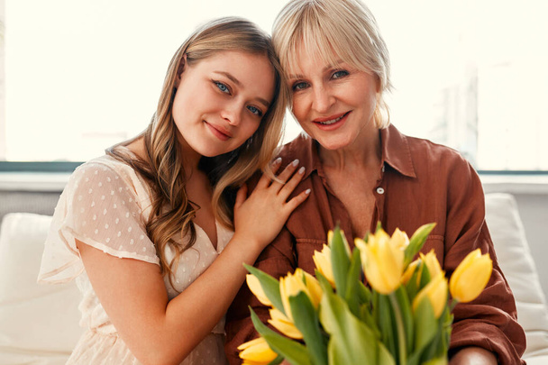 Feliz Día de la Madre y Día de la Mujer. Encantadora madre e hija con un ramo de tulipanes en la sala de estar cerca de la ventana. Amor y cuidado de la madre. - Foto, imagen
