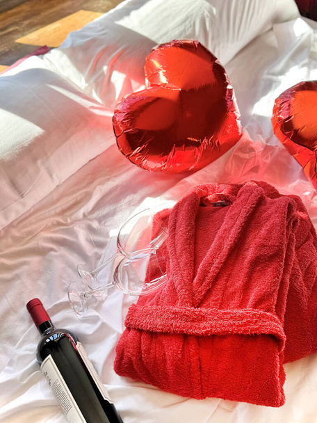 Δύο ποτήρια με μπουκάλι κόκκινο κρασί, terry κόκκινο ρόμπες και δύο μπαλόνια σε σχήμα καρδιάς σε θολή εστίαση είναι σε ένα λευκό κρεβάτι φωτίζεται από φωτεινό φως το πρωί. Κάθετη εικόνα. Ημέρα του Αγίου Βαλεντίνου. - Φωτογραφία, εικόνα