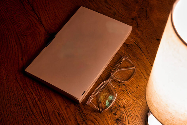 ένα λευκό-καλυμμένο βιβλίο, κομψά γυαλιά, απαλά φωτισμένο από επιτραπέζιο φωτιστικό σε ένα όμορφο φόντο δρύινο τραπέζι - Φωτογραφία, εικόνα