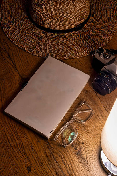 ένα γραφείο εξερευνητή, διακοσμημένο με vintage θησαυρούς. Ένα κλασικό καπέλο, διαχρονικά γυαλιά, ένα παλαιωμένο βιβλίο, και μια vintage κάμερα είναι όμορφα τοποθετημένα, λουσμένα στη ζεστή λάμψη μιας επιτραπέζιας λάμπας - Φωτογραφία, εικόνα