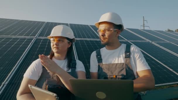Sert şapkalı ve dizüstü bilgisayarlı bir erkek ve kadın dışarıdaki güneş panellerinin arka planına karşı çalışma görevlerini tartışıyorlar. Yatırımcıyla konuşan kadın çevre mühendisi. Yeşil elektrik kavramı. - Video, Çekim