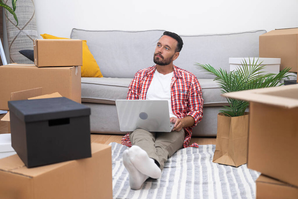 Serious male hires service to deliver furniture to new contemporary house in countryside sitting with device on floor among collected cardboard boxes of various sizes - Photo, Image