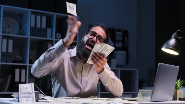 Glücklicher indischer Geschäftsmann, der Geld herumwirft, am Schreibtisch im Nachtbüro sitzt. Mann genießt Bargeld, feiert Preis, Jackpot, Glücksspielgewinn oder Gehalt. - Filmmaterial, Video