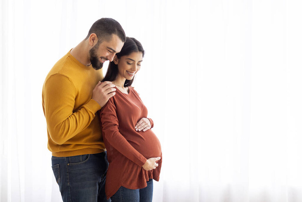 Liebender junger Ehemann umarmt seine schwangere muslimische Frau von hinten, während sie zusammen am Fenster zu Hause stehen, glückliche erwartungsvolle kaukasische Familie genießt zukünftige Elternschaft, Kopierraum - Foto, Bild