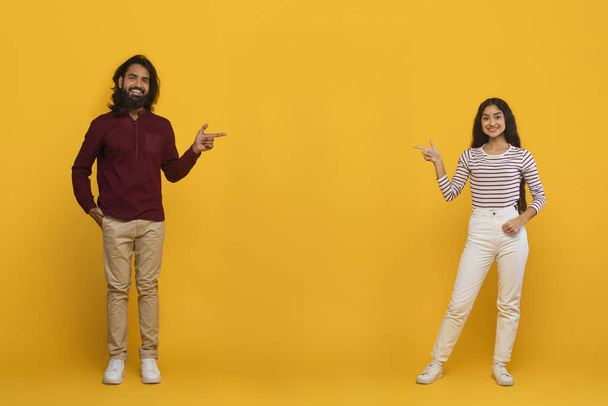 Wesoły młody Indianin mężczyzna i kobieta wskazując na przestrzeń do kopiowania tekstu lub reklamy między nimi, pokazując ładną ofertę lub ofertę dla dwojga, odizolowany na żółtym tle studio, pełna długość - Zdjęcie, obraz