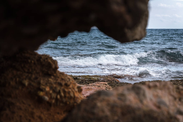 Θέα ανάμεσα στα βράχια των κυμάτων που φθάνουν στην ακτή σε μια μοναχική βραχώδη παραλία της Μεσογείου στη Σιέρα ντε Ιρτά Φυσικό Πάρκο κάτω από ένα συννεφιασμένο και θυελλώδη ουρανό, Castellon, Ισπανία - Φωτογραφία, εικόνα
