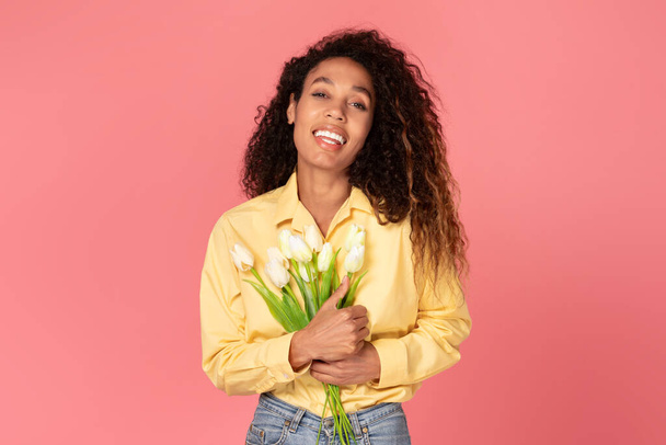 Radiante mulher negra vigas com felicidade, posando com buquê de tulipas brancas frescas contra fundo rosa encantador, sorrindo para a câmera - Foto, Imagem