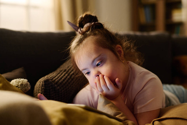 Adorabile ragazza con sindrome di Down sdraiata sul divano e tenere la mano per bocca mentre guardava cartoni animati online sullo schermo del tablet - Foto, immagini