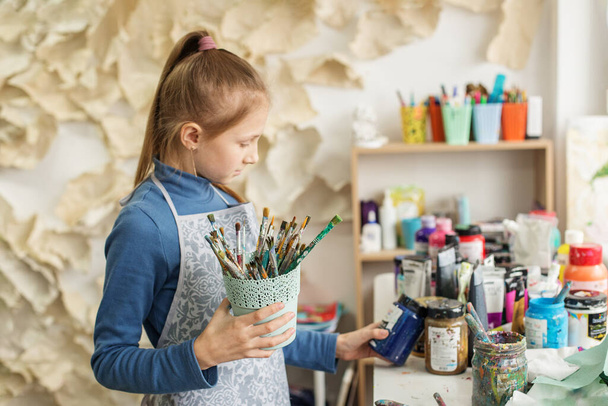 Chica joven concentrada selecciona el pincel, listo para crear, en medio de una vibrante variedad de pinturas y herramientas de arte. - Foto, Imagen