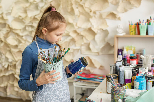 Chica joven concentrada selecciona el pincel, listo para crear, en medio de una vibrante variedad de pinturas y herramientas de arte - Foto, Imagen