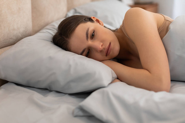 Jonge depressieve vrouw rust haar hoofd op het kussen, liggend op haar kant met een blik van zorg, reflecterende moment van zorg of diepe gedachte in bed - Foto, afbeelding