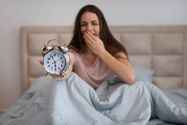 Mujer joven soñolienta bosteza mientras se sienta en la cama, presentando el reloj despertador clásico a la cámara, ilustrando la lucha de un despertar temprano, por la mañana - Foto, imagen
