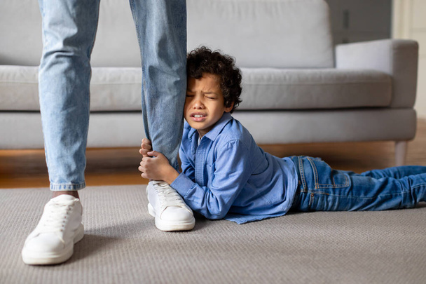 Δυσάρεστο μαύρο αγόρι που κρατά σφιχτά το πόδι του πατέρα του, δείχνοντας έκκληση για προσοχή ή άρνηση να αφήσει να πάει, ενώ ο γονέας στέκεται σταθερή, εσωτερικό σπίτι - Φωτογραφία, εικόνα
