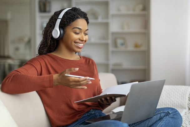 Videopuhelu. Hymyilevä musta nainen, jolla on kuulokkeet, puhelinneuvottelu kotona kannettavalla tietokoneella, onnellinen afroamerikkalainen nainen puhuu ja elehtii tietokoneen verkkokamerassa istuessaan sohvalla, vapaa tila - Valokuva, kuva