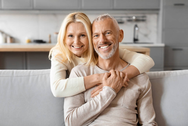 Loving beautiful blonde caucasian elderly woman hugging her cheerful handsome husband from behind, cozy home interior. Happily married senior couple enjoying time together, embracing, closeup - Photo, Image