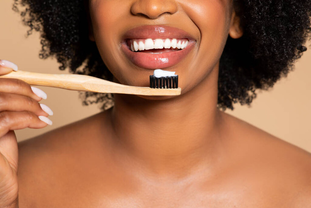 Visão de perto da mulher negra sorridente radiante prestes a escovar os dentes com escova de dentes de bambu, promovendo a higiene dental no fundo bege - Foto, Imagem