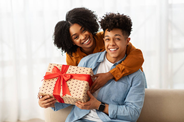 Una giovane coppia afroamericana allegra in abbigliamento casual condivide un momento gioioso mentre tiene in mano un grande regalo avvolto in carta a fantasia di cuore con un vibrante fiocco rosso a casa. - Foto, immagini