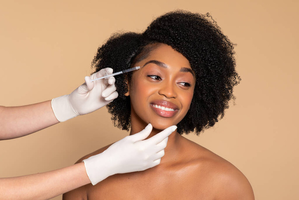 Lächelnde junge schwarze Frau erhält professionelle kosmetische Behandlung mit Spritze und demonstriert Schönheitsoperationen vor beigem Hintergrund - Foto, Bild