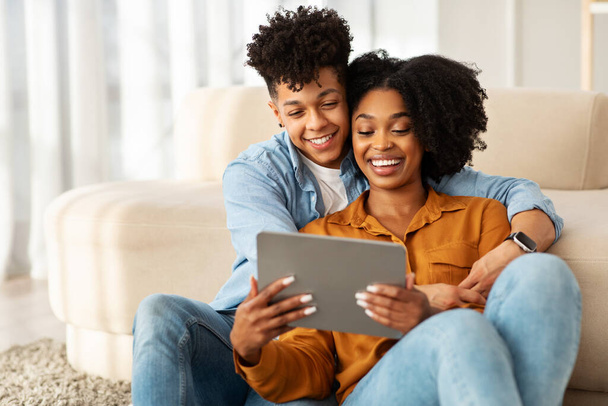 Összebújva a kanapén, az afro-amerikai fiatal pár alkalmi viseletben osztozik egy gyengéd pillanaton, miközben boldogan néznek együtt egy táblagépre egy kényelmes, világos nappaliban. - Fotó, kép