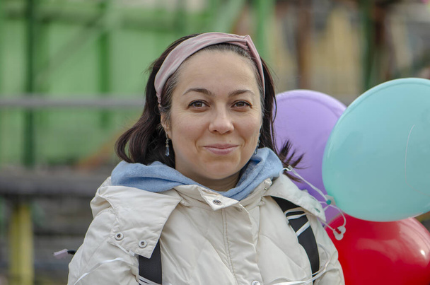 Retrato callejero de una mujer de 40-45 años de pelo oscuro sosteniendo globos multicolores sobre un fondo urbano borroso. - Foto, imagen