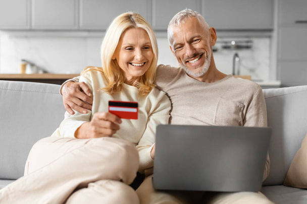 Улыбаясь привлекательный кавказский пенсионер мужчина и женщина делают покупки онлайн вместе, используя ноутбук компьютер и красную пластиковую банковскую кредитную карту, сидя дома на диване. Розничная торговля, электронная торговля - Фото, изображение