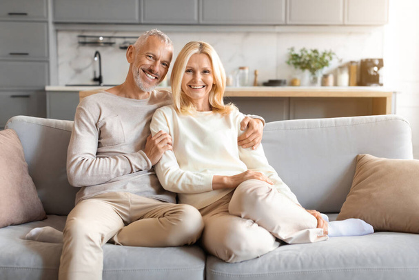 Happy loving european senior spouses sitting on couch at home modern studio apartment, embracing and smiling at camera, enjoying time together, copy space. Love, affection, family, marriage - Photo, Image