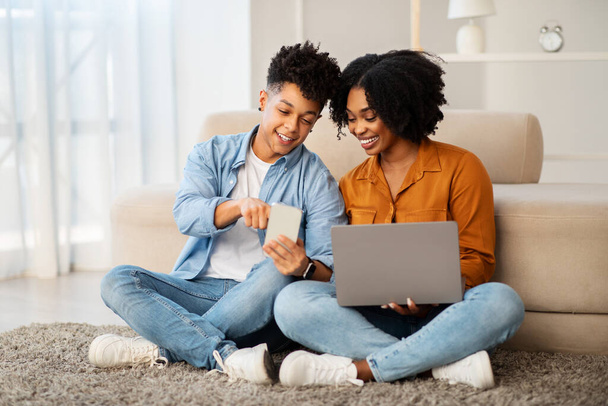 A young african american couple engages with technology in a bright living space, sharing a smile over a smartphone with a laptop open, showcasing a modern, connected lifestyle - Photo, Image