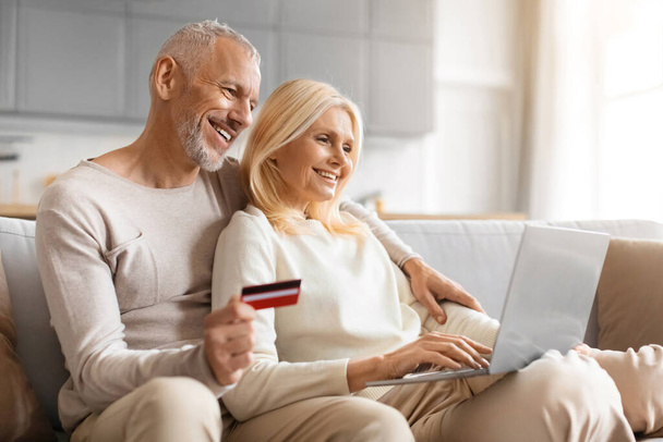 Glückliche Senioren mit Laptop und Kreditkarte, die es sich auf der Couch in der gemütlichen Studiowohnung gemütlich machen, online einkaufen oder Banking, Waren und Dienstleistungen von zu Hause aus bezahlen, Kopierraum - Foto, Bild
