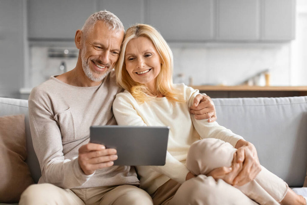 Volwassen mensen en gadgets. Happy Senior Couple met behulp van digitale tablet samen Browsing Internet Zittend op de bank thuis, Copy Space. Gepensioneerde man en vrouw lezen online nieuws op pad op weekend - Foto, afbeelding