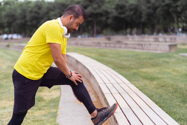 urban park, een flexibele man in zwarte legging leunt op een bank, het doen van oefeningen om zijn beenspieren te rekken.  - Foto, afbeelding